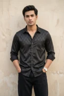 Man wearing a black satin party wear shirt with tone-on-tone jacquard check weave, glossy finish, spread collar, dark buttons, chest pocket, and full sleeves rolled at the cuffs, paired with black trousers and a silver watch