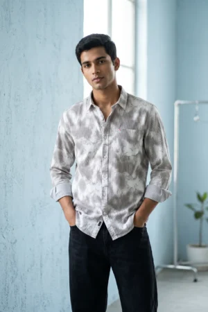 Man wearing a twill 3/1 cotton printed shirt with grey and white cloud-wash abstract print on a fine stripe base, spread collar, full sleeves rolled at the cuffs, and black trousers