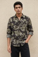 Man wearing a cotton dobby floral printed casual shirt in charcoal black with large-scale olive and cream botanical flower print, spread collar, contrast cuff lining, chest patch pocket with logo badge, and full sleeves rolled at the cuffs