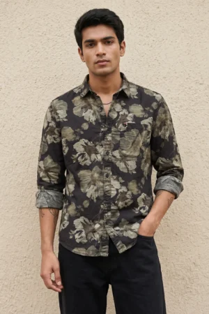 Man wearing a cotton dobby floral printed casual shirt in charcoal black with large-scale olive and cream botanical flower print, spread collar, contrast cuff lining, chest patch pocket with logo badge, and full sleeves rolled at the cuffs