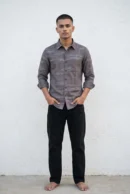 Man wearing a mocha brown natural linen shirt with all-over grey-blue thread embroidery in an abstract horizontal dash and dot pattern, spread collar, tonal buttons, chest pocket, and full sleeves rolled at the cuffs, paired with black jeans