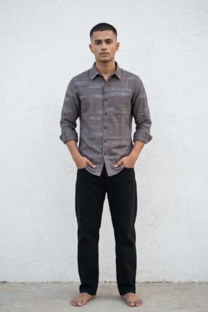 Man wearing a mocha brown natural linen shirt with all-over grey-blue thread embroidery in an abstract horizontal dash and dot pattern, spread collar, tonal buttons, chest pocket, and full sleeves rolled at the cuffs, paired with black jeans